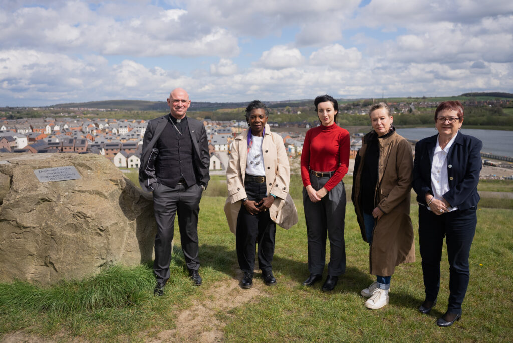 Chair and Panel members (listed left to right as Bishop Pete, Wendy Williams CBE, Dr Joanna Gilmore, Dr Angie Sutton‑Vane and Dame Mary Bousted) stand on the grassy site of Orgreave beside a stone memorial, with housing and countryside in the background.