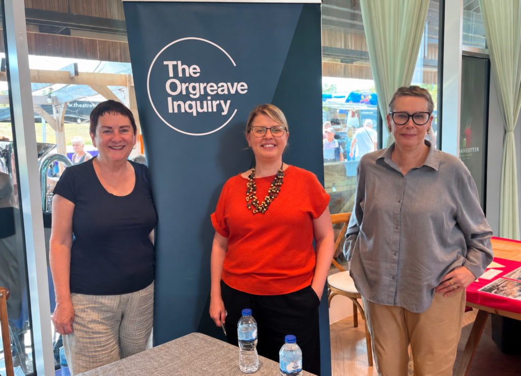 Group photograph of Panel Member Baroness Mary Bousted, Stakeholder Engagement Lead Liz Rickett and Panel Member Dr Angie Sutton-Vane standing next to an Orgreave Inquiry banner at a stakeholder engagement event. The group is positioned indoors beside large windows